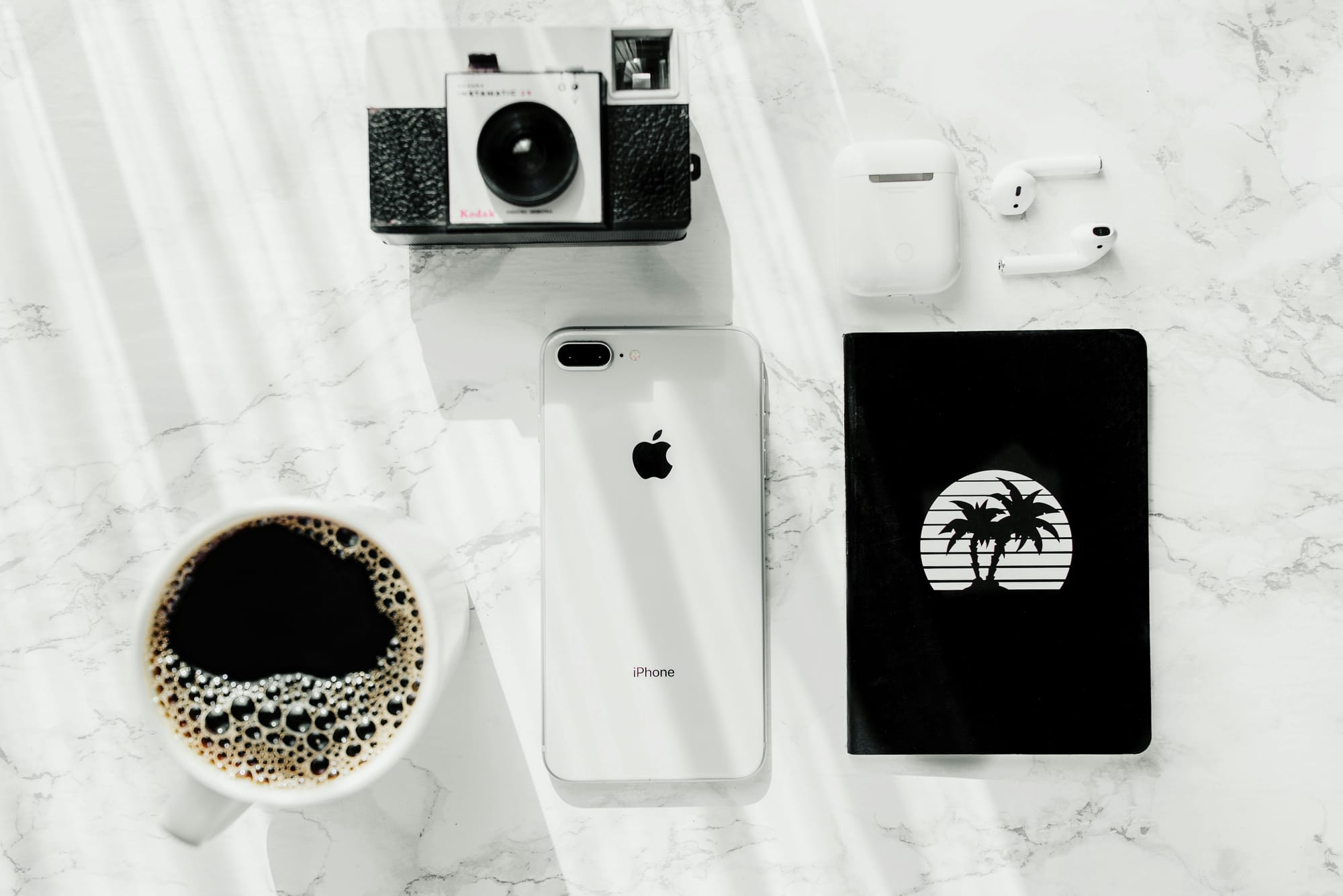 An iPhone surrounded by a cup of coffee, a camera, AirPods, and a notebook.